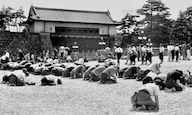 東京の歴史的な場所で、地面にひざまずいている人々の集団。背景には伝統的な建物と観衆が見える白黒写真。