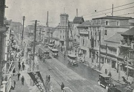 昔の日本の繁華街の風景。道路には馬車や自動車が走り、通行人が行き交っている。建物や電柱が並んでいる。