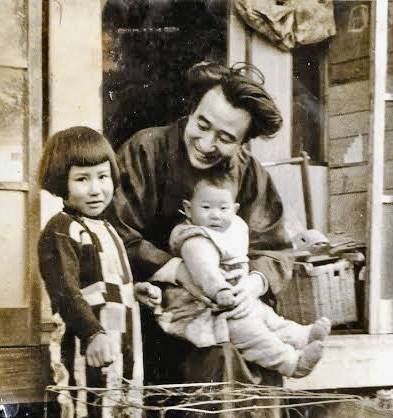 母親と二人の子供が一緒にいる古い写真。左側の子供はストライプの服を着ており、右側の母親は赤ちゃんを抱いて笑顔を見せている。