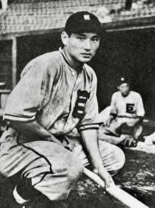 野球選手がバットを持って座っているモノクロの写真。背景には他の選手が見える体育館のような環境。