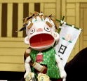 緑の衣装を着た人形が大きな口を開けている。背景には伝統的な舞台があり、白い旗が立てられている。